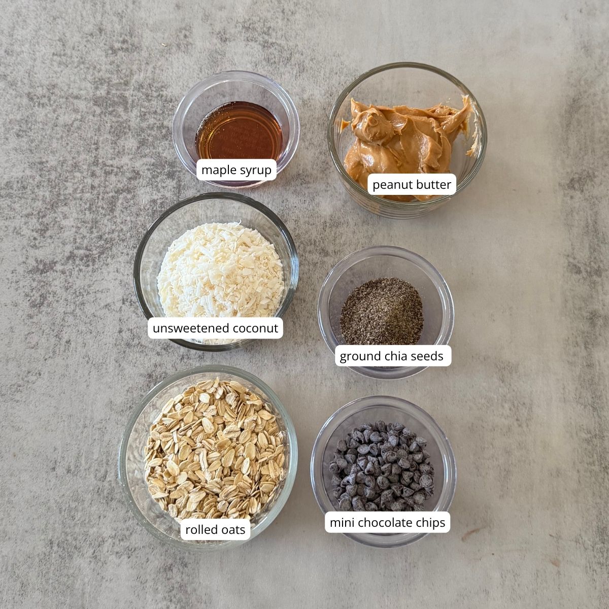 Ingredients for no-bake chocolate coconut oat bars, including peanut butter, maple syrup, oats, ground chia seeds or flaxseed, shredded coconut, and mini chocolate chips.